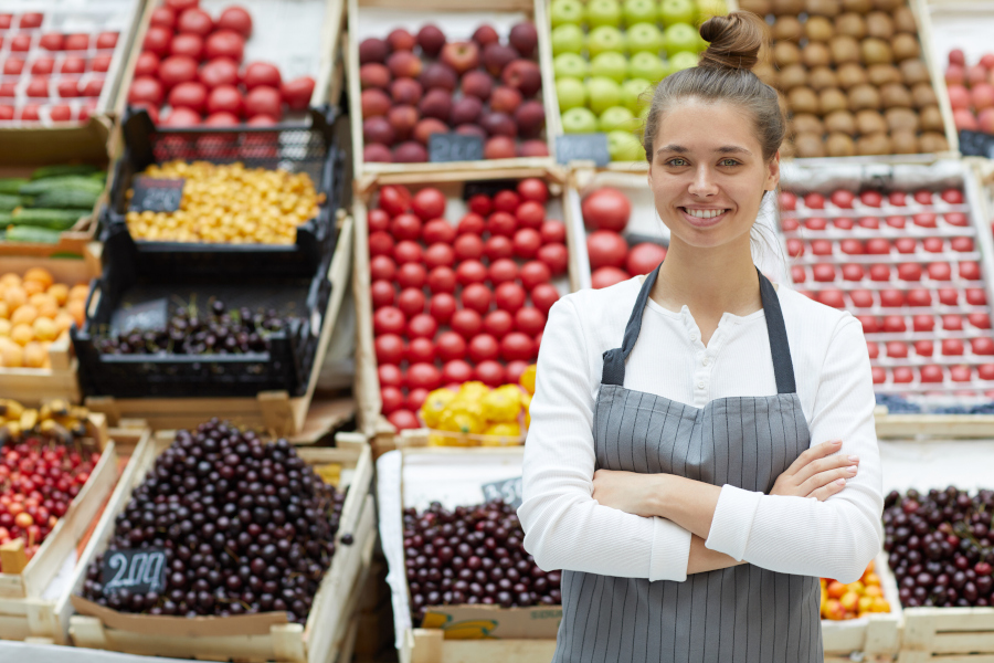 Junge Verkäuferin mit Schürze steht lächelnd vor einer Auslage mit frischem Obst und Gemüse.