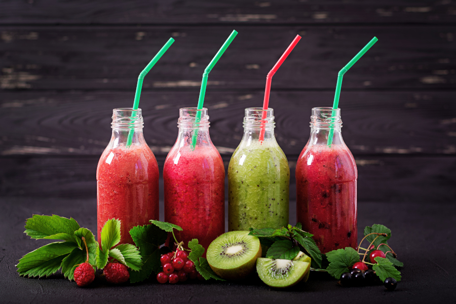 Vier Smoothies in Glasflaschen mit Strohhalmen, davor Beeren und aufgeschnittene Kiwi.