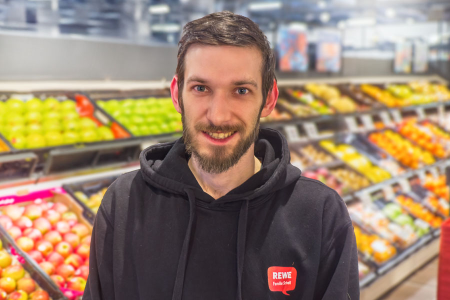 Mann im schwarzen Hoodie lächelt vor einer Obst- und Gemüseauslage im Supermarkt.