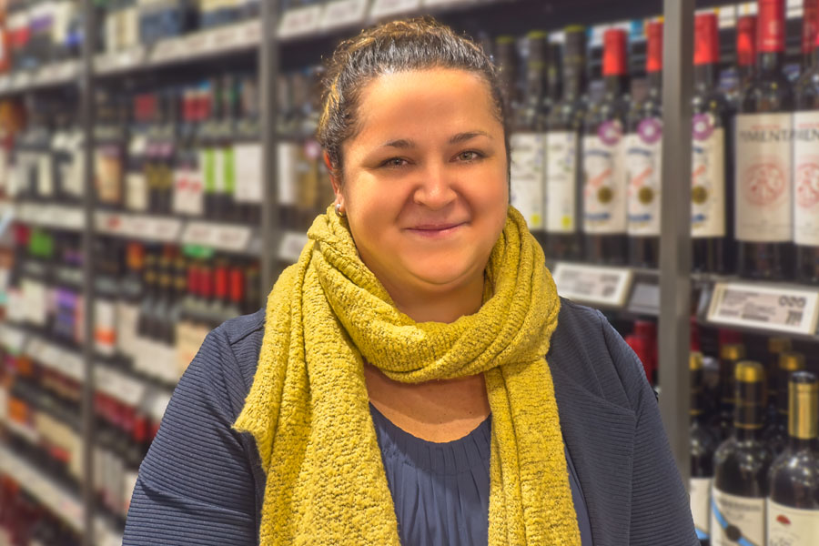 Frau mit gelbem Schal lächelt vor einem Weinregal.