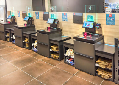 Mehrere Self-Checkout-Kassen im Supermarkt mit Touchscreens und Ablagen für Einkaufstüten.