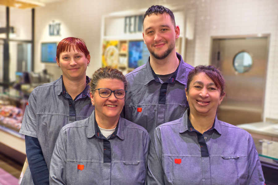 Vier Mitarbeitende in grauen Arbeitshemden lächeln für ein Gruppenfoto in einem Laden.